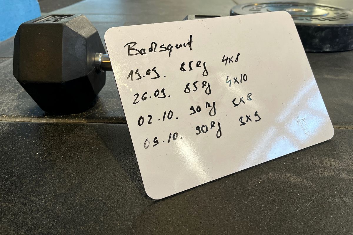 Whiteboard mit handschriftlichen Trainingseinträgen für Backsquat neben einer Kurzhantel auf dem Boden einer CrossFit-Box